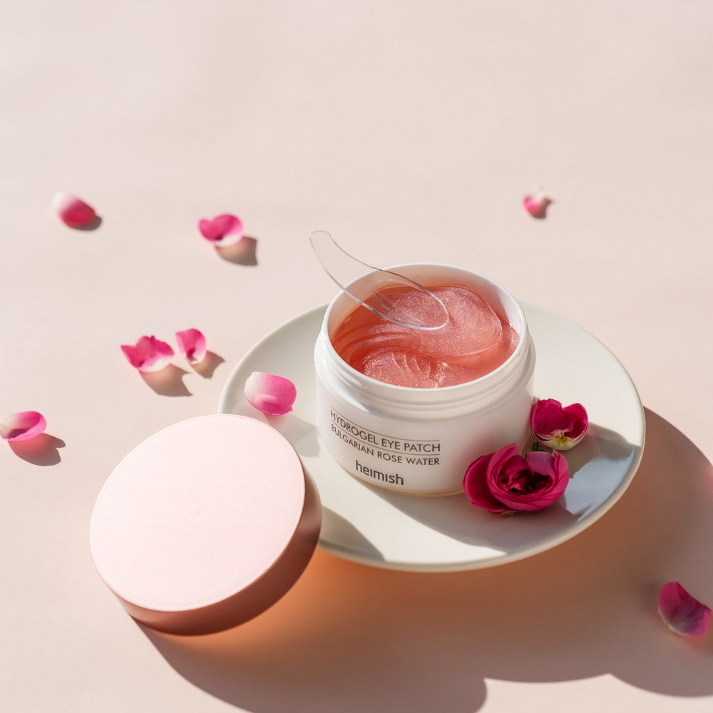 A jar of Heimish Bulgarian Rose Hydrogel Eye Patches with a pink cream inside, surrounded by scattered rose petals and a spoon for application.